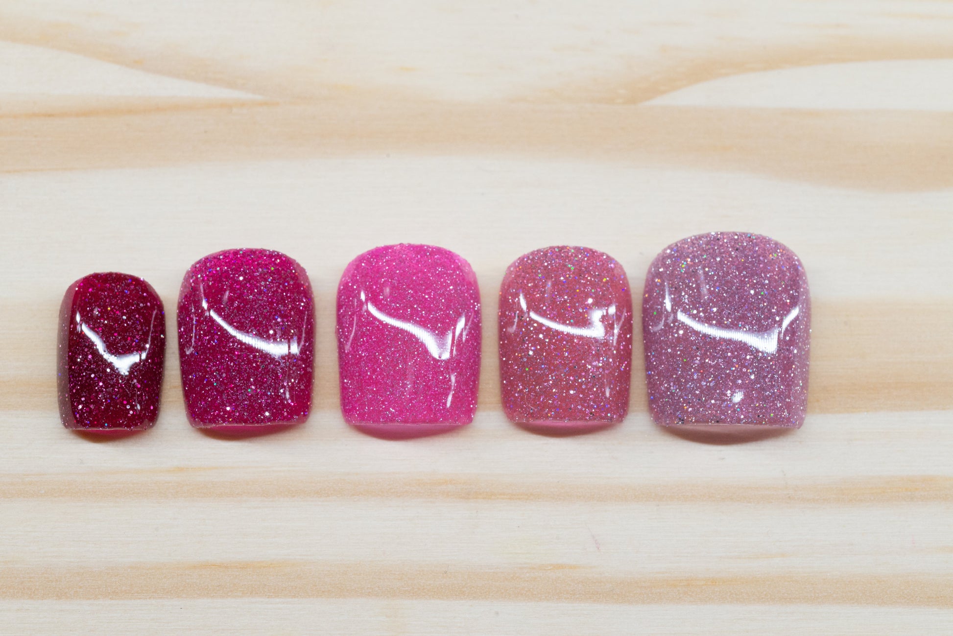 Pink glittery press on nails that range from dark to light across the fingers featured on a wooden background