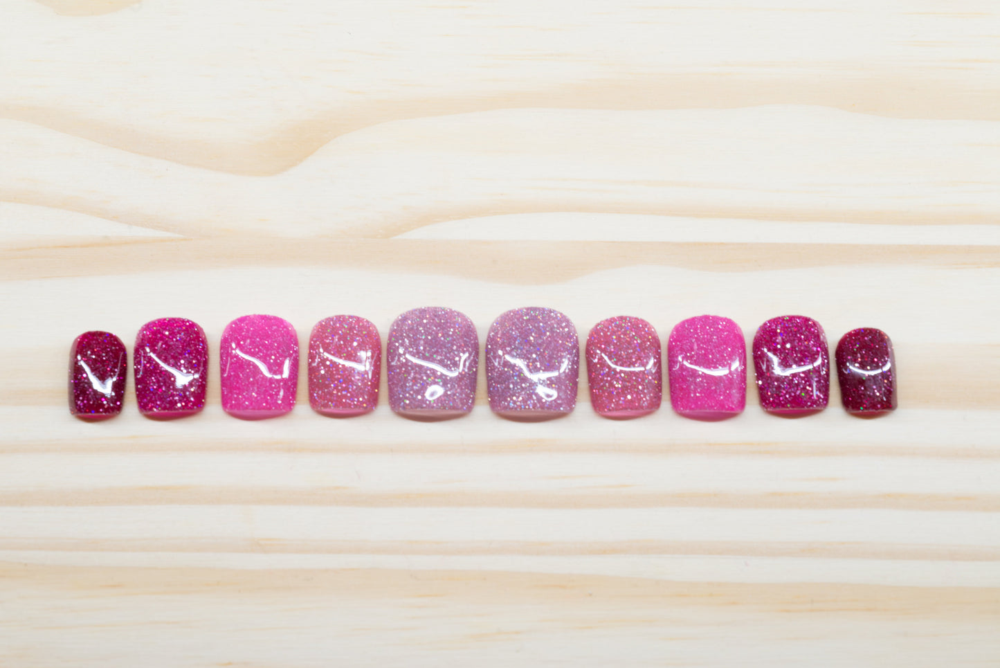 Pink glittery press on nails that range from dark to light across the fingers featured on a wooden background