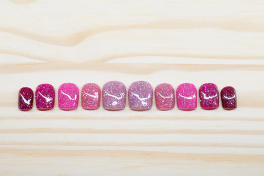 Pink glittery press on nails that range from dark to light across the fingers featured on a wooden background